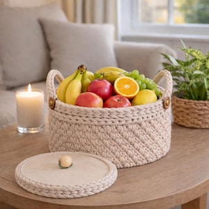 Peut inclure: Un panier beige tricoté à la main rempli de fruits frais, dont des bananes, des pommes, des raisins et une orange. Le panier a deux poignées et est posé sur une table en bois. Un couvercle assorti est à côté. Une bougie allumée et une plante sont également visibles.
