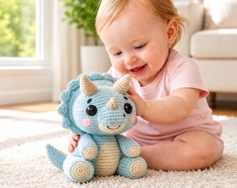 May include: A light blue crocheted triceratops plush toy with tan horns and belly. The dinosaur has large black eyes, pink cheeks, and a smiling mouth. The toy is sitting on a white carpet.