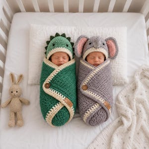 May include: Two infants wrapped in crochet swaddles, one green with dinosaur spikes and one gray with elephant ears. Both swaddles feature cream-colored trim, a single wooden button closure, and a small rectangular brand tag on the front.