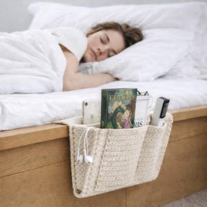 Op de afbeelding: Een crèmekleurige, gehaakte bedorganizer met drie vakken, hangend aan een houten bedframe. De vakken bevatten een smartphone, een boek met de titel "Fairy Tales", een afstandsbediening en oortelefoons. Op de achtergrond slaapt een persoon.