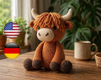 May include: A crocheted Highland cow plush toy in shades of brown and tan. The handmade stuffed animal has a shaggy mane, small horns, and a light-colored muzzle. The cow is sitting on a wooden surface, with a mug and plants in the background.