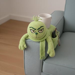 May include: A light green crocheted Grinch-themed couch armrest organizer. The organizer has a face with embroidered details, arms, and a scarf. A white mug, remote control, and glasses are in the pockets.