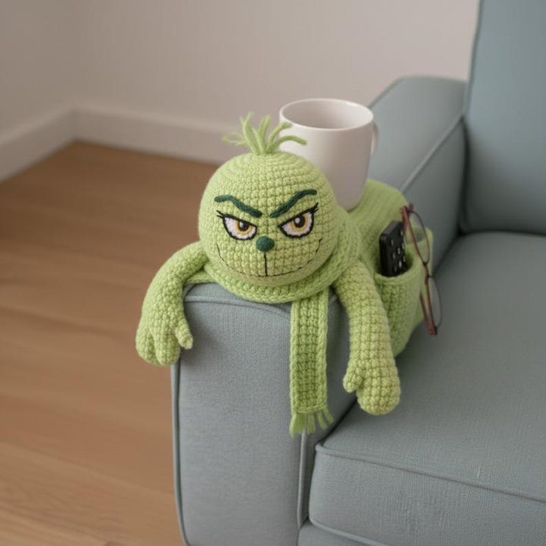 May include: A light green crocheted Grinch-themed couch armrest organizer. The organizer has a face with embroidered details, arms, and a scarf. A white mug, remote control, and glasses are in the pockets.