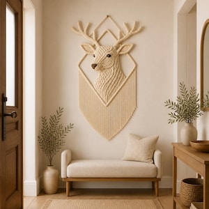 Op de afbeelding: Een beige macramé wanddecoratie van een hertenkop met een geometrisch ontwerp. De hertenkop is gecentreerd in een ruitvorm, met franjes aan de onderkant. De kamer heeft ook een bank, planten en een houten tafel.