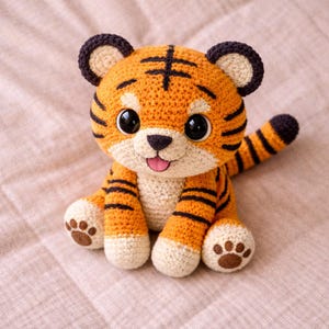 Può includere: Un peluche a forma di tigre all'uncinetto con strisce arancioni e nere, grandi occhi neri e una lingua rosa. La tigre fatta a mano ha zampe e coda color crema ed è seduta su una superficie rosa.