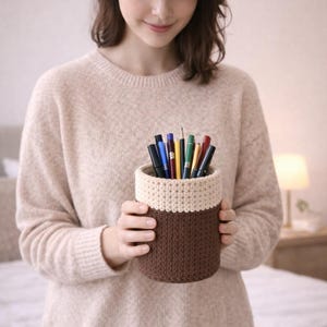 May include: A handmade crochet pen holder in brown and cream colors. The cylindrical container is filled with various pens and pencils. The holder is being held by a person wearing a beige sweater.