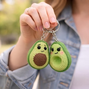 Puede incluir: Dos llaveros de aguacate de ganchillo. Cada aguacate es verde con un hueso marrón y una cara sonriente. Un llavero tiene pestañas y rubor rosa. Los llaveros están unidos a un llavero plateado.