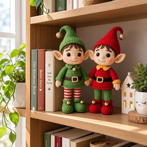 May include: Two crocheted elf dolls standing on a shelf. One elf wears a green outfit with a matching hat, and the other wears a red outfit with a matching hat. Both dolls feature pointed ears, rosy cheeks, brown hair, and small gold bells on their hats and shoes.