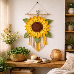 May include: A handcrafted wall hanging featuring a large, crocheted sunflower. The sunflower has yellow petals, a brown center, and green leaves. It is suspended from a wooden dowel with cream-colored macrame and yellow tassels, creating a rustic, decorative piece.