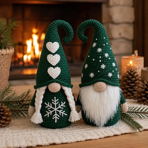 May include: Two handmade crochet gnomes in dark green. The left gnome features three white hearts on its tall hat, a large white snowflake on its body, and two white braided pigtails. The right gnome has white snowflake embroidery and a long white beard.