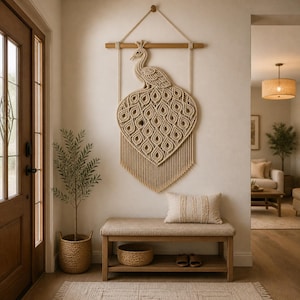 May include: A large macrame peacock wall hanging in natural tones is displayed above a wooden bench. The peacock design features a detailed body and tail, with a fringe at the bottom. A woven basket and a small tree in a basket are also in the image.