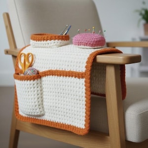 May include: A white and orange crochet armrest organizer with pockets, a small basket, and a pink pincushion. The organizer is draped over a wooden chair arm, holding scissors and yarn. The design features a textured stitch and a contrasting orange border.