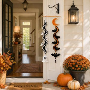 May include: A hanging wall decoration featuring two crocheted spiral streamers suspended from a wooden dowel. One streamer is black and white with small ghost figures, while the other is black and orange with bats and a crescent moon.