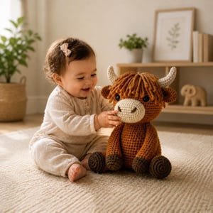 May include: A crocheted Highland cow plush toy with brown and tan yarn. The toy has a cream-colored muzzle, white horns, and dark brown eyes. The plush is sitting on a beige carpet.