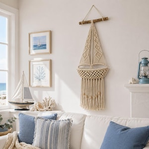 May include: A cream-colored macrame wall hanging shaped like a sailboat, suspended from a horizontal wooden dowel. The design features intricate knotted patterns forming the sails and hull, finished with long fringe and small wooden beads at the base.