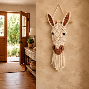 Puede incluir: Un colgante de pared con cabeza de burro hecho con macramé. El burro tiene la cara color crema con orejas y hocico marrones. Ojos de botón negros y una melena de flecos completan el diseño. Colgado de una varilla de madera.