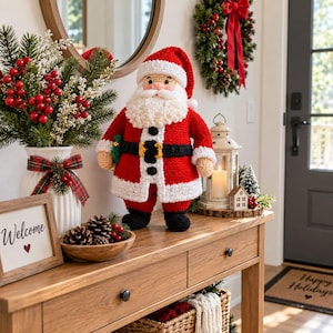 May include: A crocheted Santa Claus figurine featuring a red coat, white trim, a black belt with a yellow buckle, and black boots. The Santa has a white beard and a red hat. It stands on a wooden console table next to a white vase with red berries.
