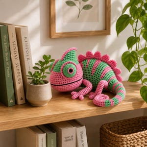 Op de afbeelding: Een gehaakt kameleon-speeltje met afwisselende horizontale strepen van felgroen en roze garen. Het knuffeldier heeft een gekrulde staart, een stekelige rugkam en een opvallend oog. Het staat op een houten plank naast een kleine plant in een pot.