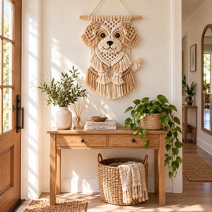 Puede incluir: Un tapiz de pared hecho de cuerda de macramé color crema y tostado, con forma de perro con orejas caídas y cola. La pieza cuelga de un soporte de madera y presenta intrincados patrones de nudos que forman la cara, el cuerpo y el pelaje.