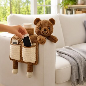 Puede incluir: Un oso de peluche marr&oacute;n tejido a ganchillo con un organizador de reposabrazos a juego. El organizador tiene dos bolsillos, un portavasos y un bolsillo para el tel&eacute;fono. El oso est&aacute; sentado en un sof&aacute; blanco. El organizador est&aacute; dise&ntilde;ado para el reposabrazos.