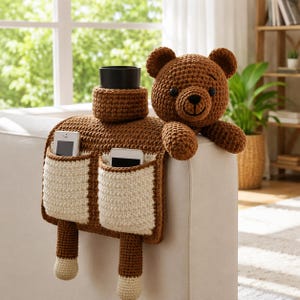 Puede incluir: Un organizador de brazo de sof&aacute; tejido a ganchillo marr&oacute;n y crema con un dise&ntilde;o de oso de peluche. Cuenta con dos bolsillos para tel&eacute;fonos, un portavasos y una cabeza de oso de peluche. La cabeza del oso es marr&oacute;n con nariz y ojos negros.