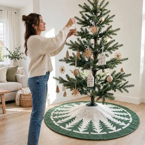 May include: A round, crocheted Christmas tree skirt featuring a repeating pattern of white pine trees on a dark green background, with a white center section. The skirt is placed at the base of a green artificial tree decorated with wooden ornaments.