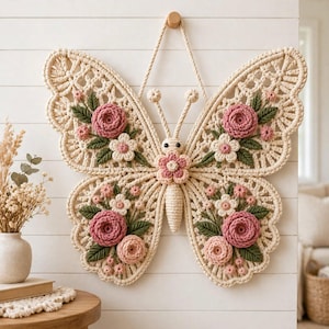 May include: A cream-colored macrame butterfly wall hanging featuring intricate knotted wing patterns. The butterfly is decorated with crocheted pink roses, smaller light pink blossoms, and dark green leaves attached to the wings and center body.