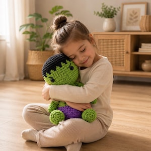 May include: A young girl embraces a handmade crochet doll. The doll is green with black hair, a purple waist, and green feet. The girl is wearing a cream-colored long-sleeved shirt and light-colored pants. The doll is approximately 12 inches tall.