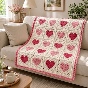 May include: A rectangular crochet blanket featuring a grid of square motifs, each containing a heart in varying shades of pink and red. The blanket is bordered with a scalloped pink edge and is constructed from off-white yarn throughout.