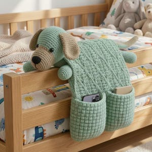 Könnte beinhalten: Ein handgefertigter, gehäkelter Hunde-Bett-Organizer in Hellgrün, Beige und Schwarz. Der hundeformige Organizer hat zwei Taschen und hängt über der Seite eines Holzbetts. Der Kopf und der Körper des Hundes sind sichtbar.