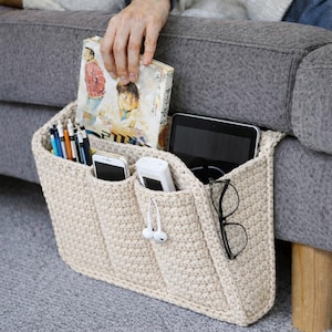 May include: A cream-colored, crocheted sofa arm organizer with multiple pockets. The organizer holds a book, pens, a smartphone, a tablet, a portable charger, and eyeglasses. The organizer is attached to the arm of a gray sofa.