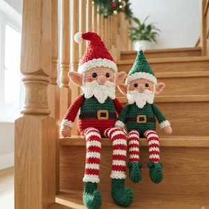 Op de afbeelding: Twee handgemaakte gehaakte elfjes met puntmutsen, witte baarden en lange bungelende benen. Het grotere elfje draagt een rood-groene outfit met witte randen, terwijl het kleinere elfje een groene outfit met witte randen draagt. Beide hebben bruine riemen met goudkleurige gespen en gestreepte rood-witte benen.