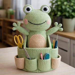 Puede incluir: Un organizador hecho a mano con forma de rana de ganchillo en verde claro, con vientre beige y mejillas rosas. La rana tiene ojos grandes y redondos y sostiene una cesta de almacenamiento con bolsillos. Se ven tijeras, bolígrafos y otros útiles.
