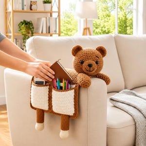 Puede incluir: Un organizador de reposabrazos de oso de peluche marr&oacute;n tejido a ganchillo con dos compartimentos con bolsillos blancos. El oso est&aacute; sentado en un brazo de sof&aacute; beige. Los bolsillos contienen un tel&eacute;fono, bol&iacute;grafos y un cuaderno marr&oacute;n.