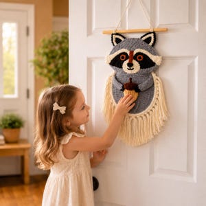 May include: A handmade crochet raccoon wall hanging. The raccoon has a gray body, black mask, and holds a tan acorn. The wall hanging has cream-colored fringe and is suspended from a wooden dowel. A young girl is reaching for the raccoon.