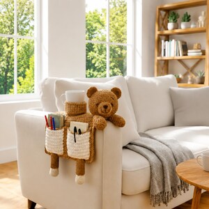 Puede incluir: Un organizador de brazo de sof&aacute; con forma de oso de peluche marr&oacute;n con dos bolsillos blancos y un portavasos. El organizador est&aacute; tejido a ganchillo y contiene bol&iacute;grafos, cuadernos y una taza blanca. Los brazos del oso descansan en el sof&aacute;.