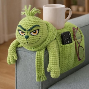 May include: A green crocheted sofa armrest featuring a Grinch character design. The armrest has a pocket for a remote and glasses, and holds a white mug. The Grinch has a smiling face with yellow eyes and a green scarf.