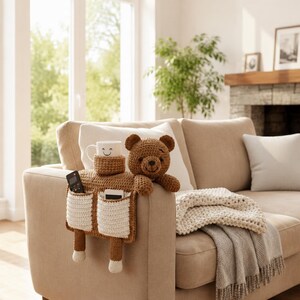 Puede incluir: Un organizador de brazo de sof&aacute; tejido a ganchillo marr&oacute;n con un dise&ntilde;o de oso de peluche. Cuenta con un portavasos, dos bolsillos y una cabeza y brazos de oso de peluche. Una taza blanca con una cara sonriente est&aacute; en el portavasos.