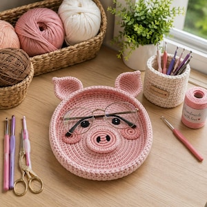 May include: A pink crocheted pig-shaped tray with two ears, a snout, and black eyes. The tray holds a pair of thin-framed glasses. Nearby are various crochet hooks, a pair of gold scissors, a small cream-colored basket, and a spool of pink yarn.