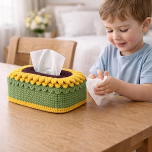 May include: A handmade, crochet tissue box cover in the shape of a sunflower. The box features a green base, yellow petals, and a brown center. White tissues are visible. The cover is on a wooden table.