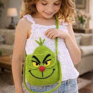 Op de afbeelding: Een groene gehaakte crossbodytas met het gezicht van de Grinch. De tas heeft een lachende mond, een rode neus en gele ogen. De riem is groen en de tas wordt over de schouder gedragen.