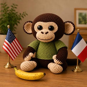 Könnte beinhalten: Gehäkelte Affenpuppe in Braun, Beige und Grün, mit einem grünen Shirt. Ein gelbe Banane und Miniatur-Flaggen von Amerika und Frankreich sind im Vordergrund. Die handgefertigte Puppe hat ein freundliches Gesicht.