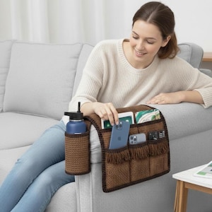 Op de afbeelding: Een bruine, gehaakte sofa-arm organizer met meerdere vakken en een waterfleshouder. De organizer bevat een blauwe waterfles, een tablet, een smartphone en andere kleine items. De franje voegt een decoratief tintje toe.