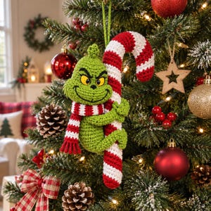 May include: A crocheted green creature ornament with yellow eyes and a mischievous expression, wearing a red and white striped scarf and holding onto a large red and white striped candy cane, suspended by a green string from a Christmas tree.