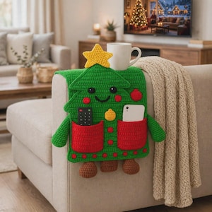 May include: A green crochet Christmas tree armrest organizer featuring a yellow star topper, two red pockets, and brown feet. The organizer has a smiling face, red cheeks, and decorative ornaments. It holds a black remote and a white smartphone.