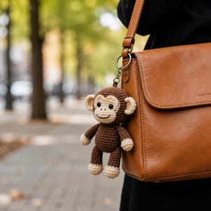May include: A crocheted monkey keychain in brown and cream colors. The monkey has a smiling face and is attached to a silver keyring. The keychain is hanging next to a brown leather purse.