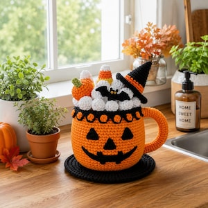 Op de afbeelding: Een gehaakte oranje pompoenmok gevuld met witte garenbolletjes, een kleine oranje pompoen, maissnoepjes, een zwarte vleermuis en een zwarte heksenhoed. De mok heeft een jack-o'-lantern gezicht en staat op een zwarte gehaakte onderzetter.