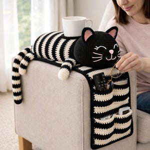 May include: A black and white striped crochet cat-shaped armrest organizer with pockets. The cat has a smiling face with pink inner ears. A white mug sits on the cat's back. The organizer is on a beige upholstered chair.