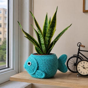 May include: A turquoise crocheted fish-shaped plant pot cover featuring a single white and black googly eye on the side, a textured fin, and a tail, containing a tall snake plant with green and yellow variegated sword-shaped leaves.