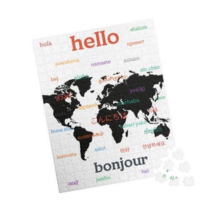 Puede incluir: Un rompecabezas que muestra un mapa mundial negro sobre un fondo blanco. La palabra hello está impresa en letras rojas grandes en la parte superior, y bonjour en letras gris oscuro grandes en la parte inferior. Varios saludos en múltiples idiomas están dispersos por el mapa en diferentes colores. Varias piezas sueltas del rompecabezas son visibles en la esquina inferior derecha.